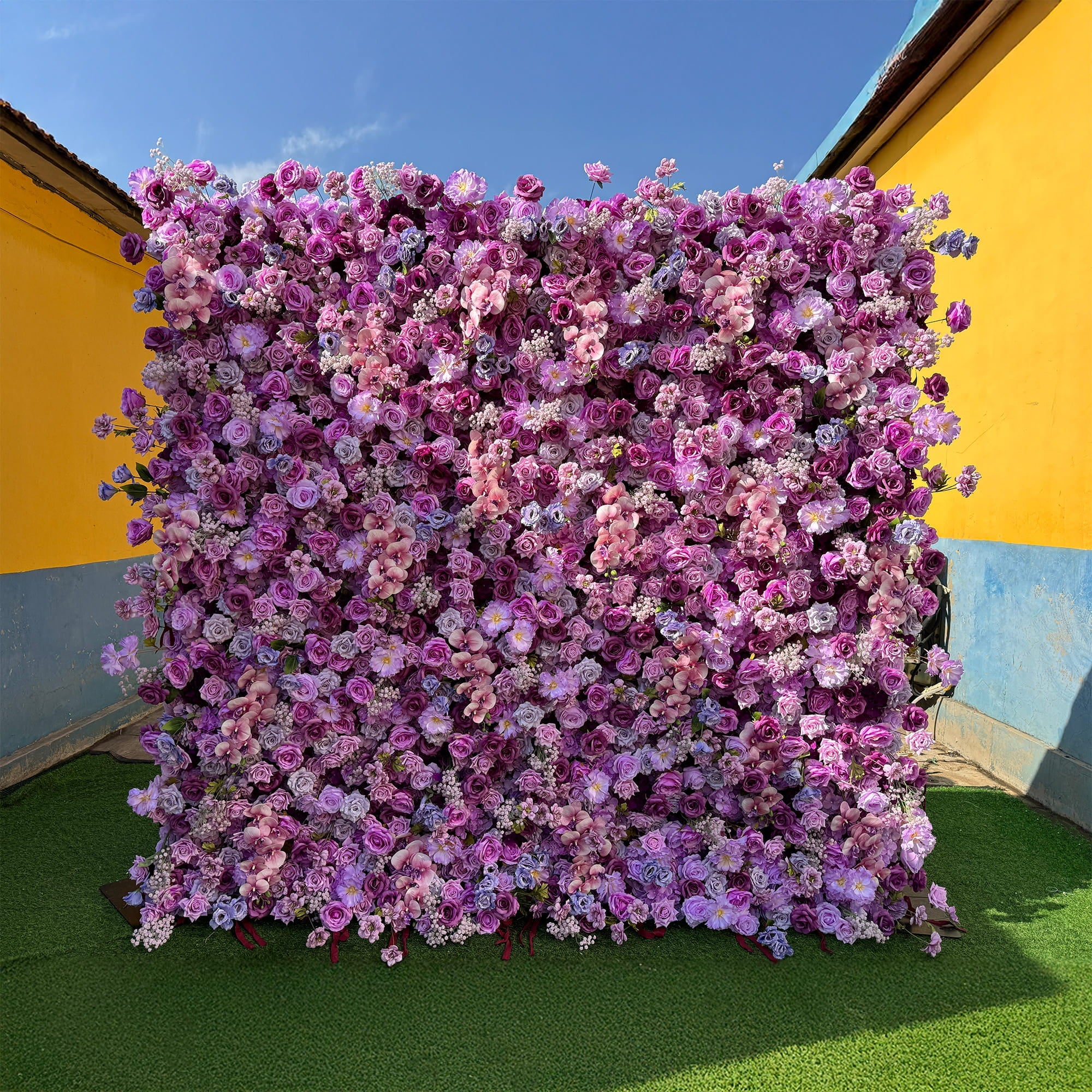 This image features a vibrant flower wall composed of artificial flowers. The wall is densely packed with roses in various shades of purple, from deep violet to soft lavender, along with hints of pink and white blooms interspersed throughout. The background consists of a yellow and blue - painted wall and a green artificial grass base. The overall style is lush and romantic, ideal for events or photo - opportunities.