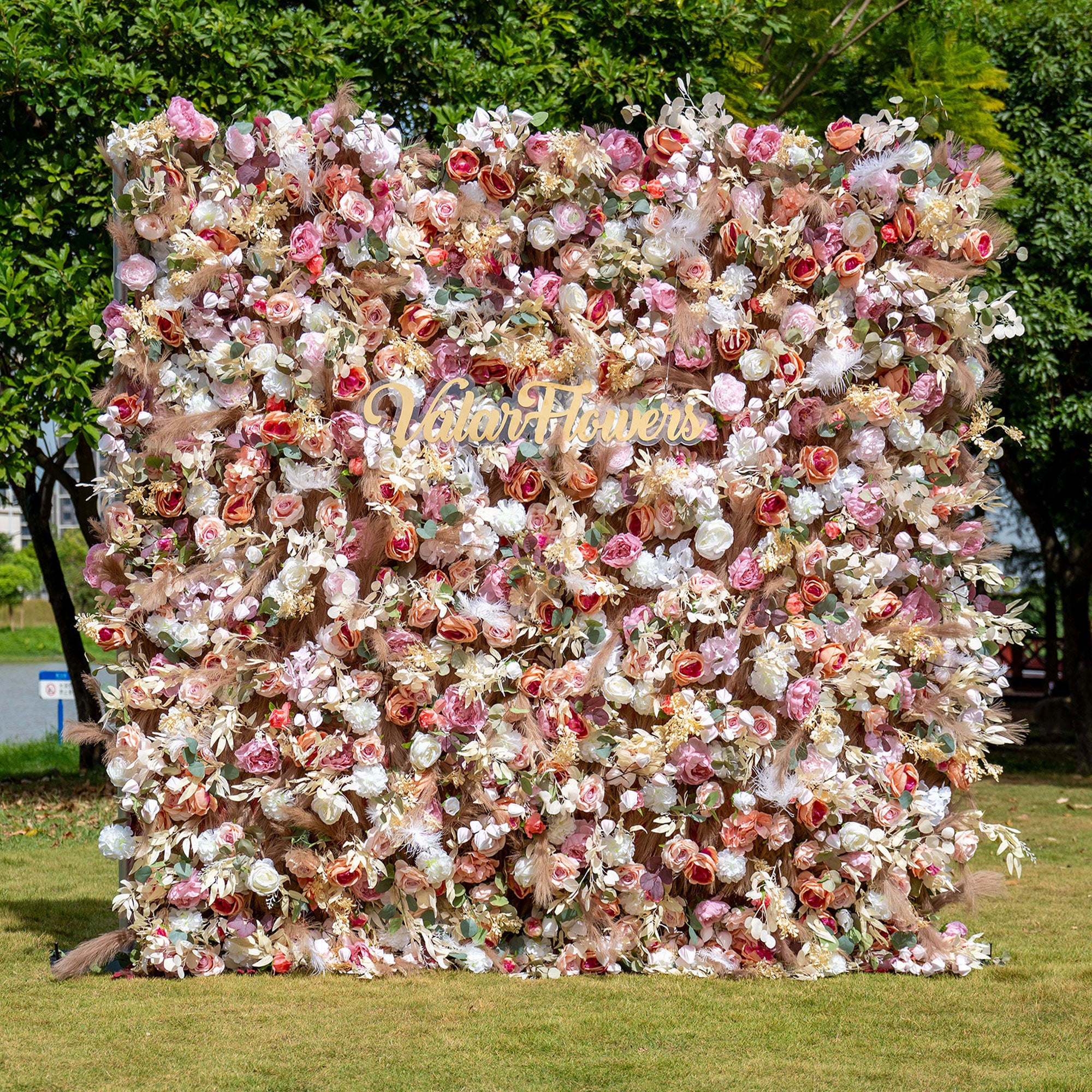An artificial flower wall with a fabric base, adorned with a variety of flowers in pink, white, and other colors, with a sign that reads 'WEDDING' in the center.