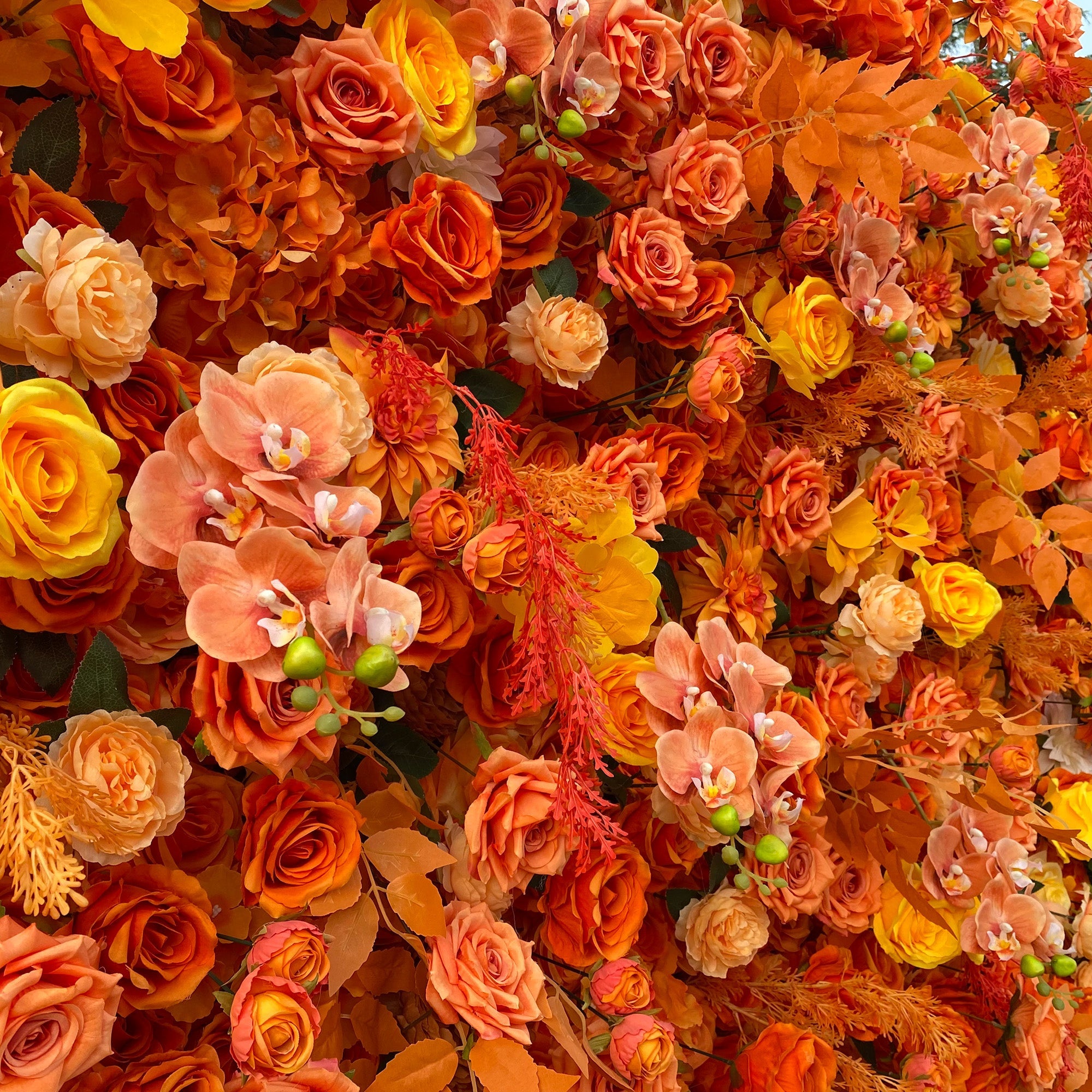 This image showcases a richly textured floral arrangement dominated by warm hues. The flowers, primarily artificial, feature an array of oranges, from deep burnt orange to soft peach, along with touches of bright yellow. Prominent flower types include roses and orchids, interspersed with decorative foliage and small green buds. The overall style is vibrant and autumnal, creating a warm and inviting visual display.
