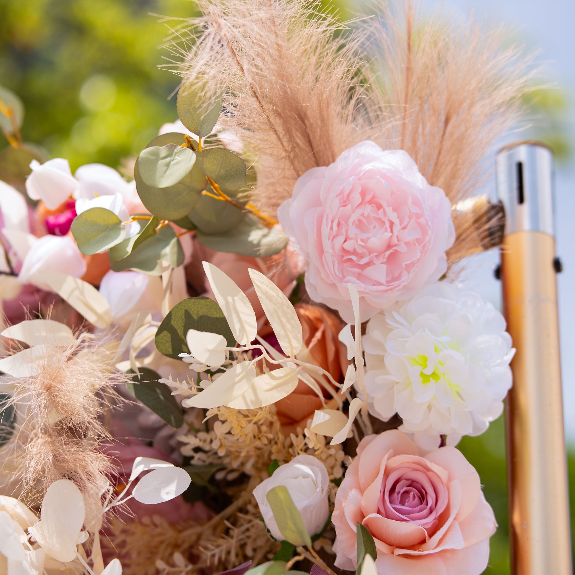Decorative floral arrangement with pink roses, green leaves, and dried pampas grass against a blurred natural background.