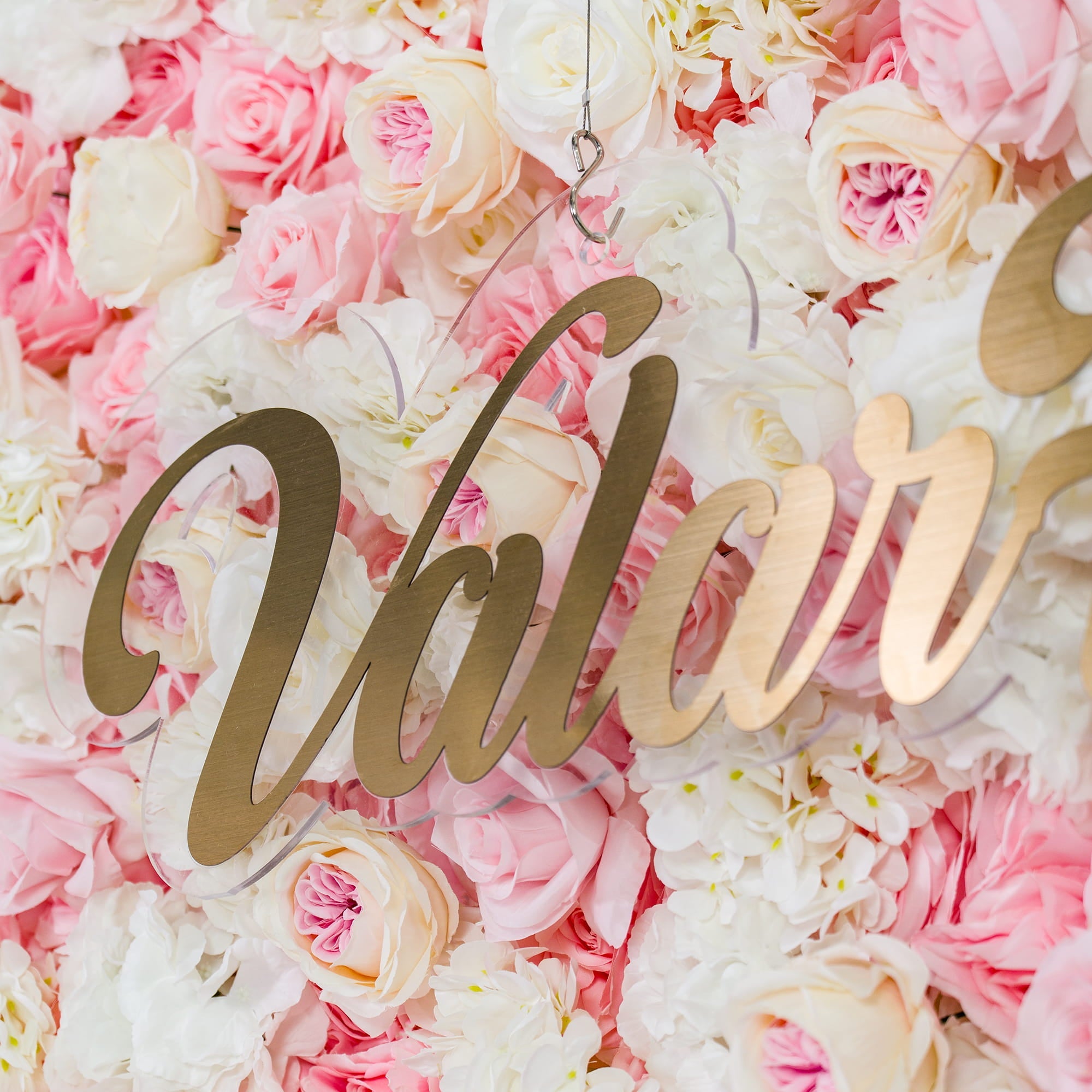 This photo showcases a lush floral backdrop with a dense arrangement of pink and white roses. The soft pastel color palette creates a romantic and elegant atmosphere. The word "Valar" in elegant gold lettering stands out prominently. Ideal for weddings or bridal showers, it exudes a dreamy and sophisticated style.