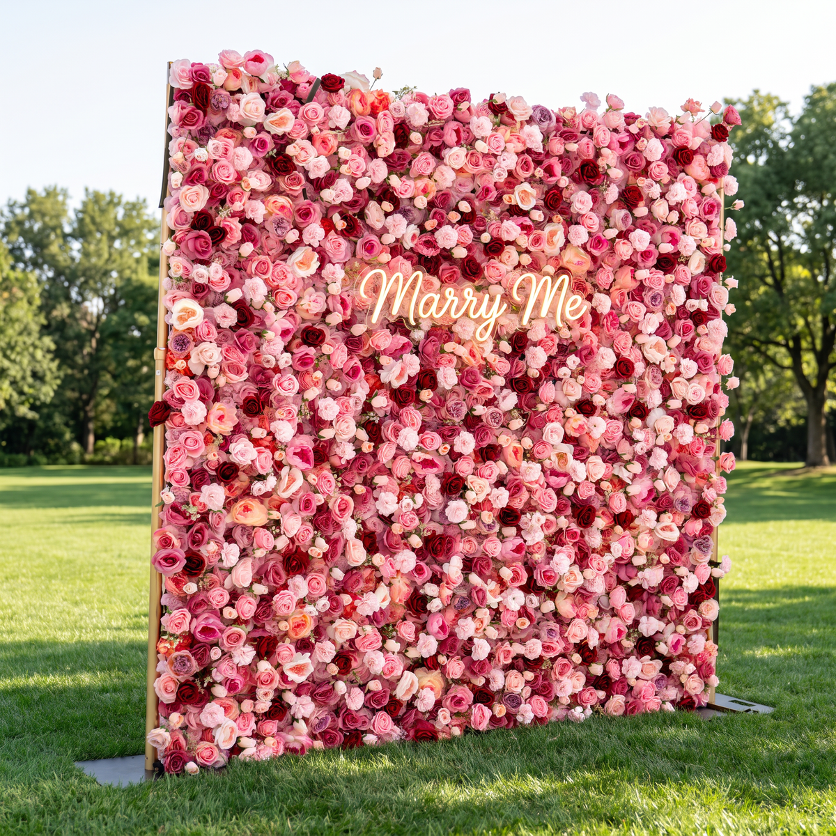 A large floral photo backdrop stand covered in pink, red, and white flowers, with a glowing 'Marry Me' sign in the center, set outdoors on a grassy lawn with trees in the background.
