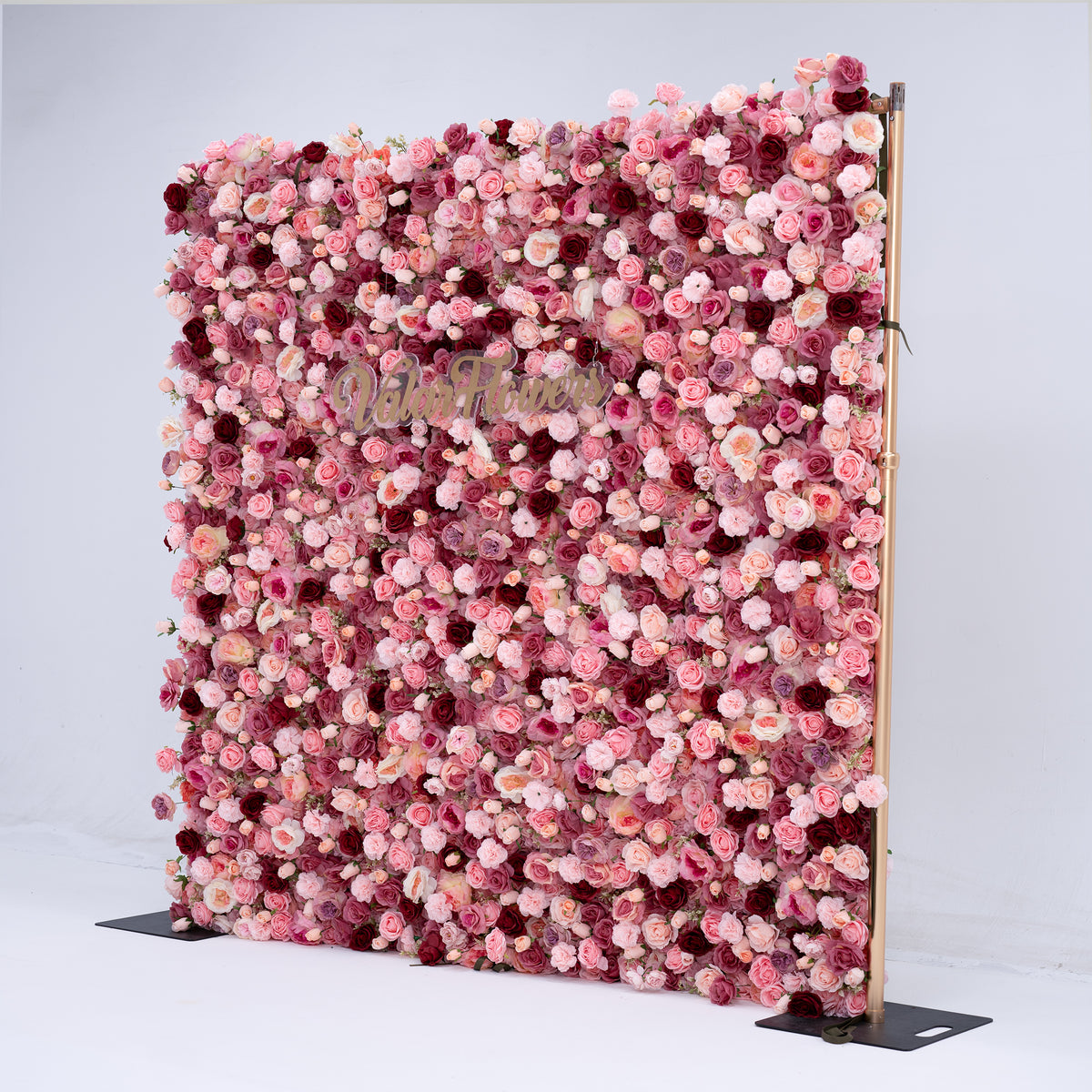 A large vertical floral backdrop wall covered in dense pink, rose, and burgundy faux roses, mounted on a thin wooden frame with black metal feet, photographed against a light gray studio background.