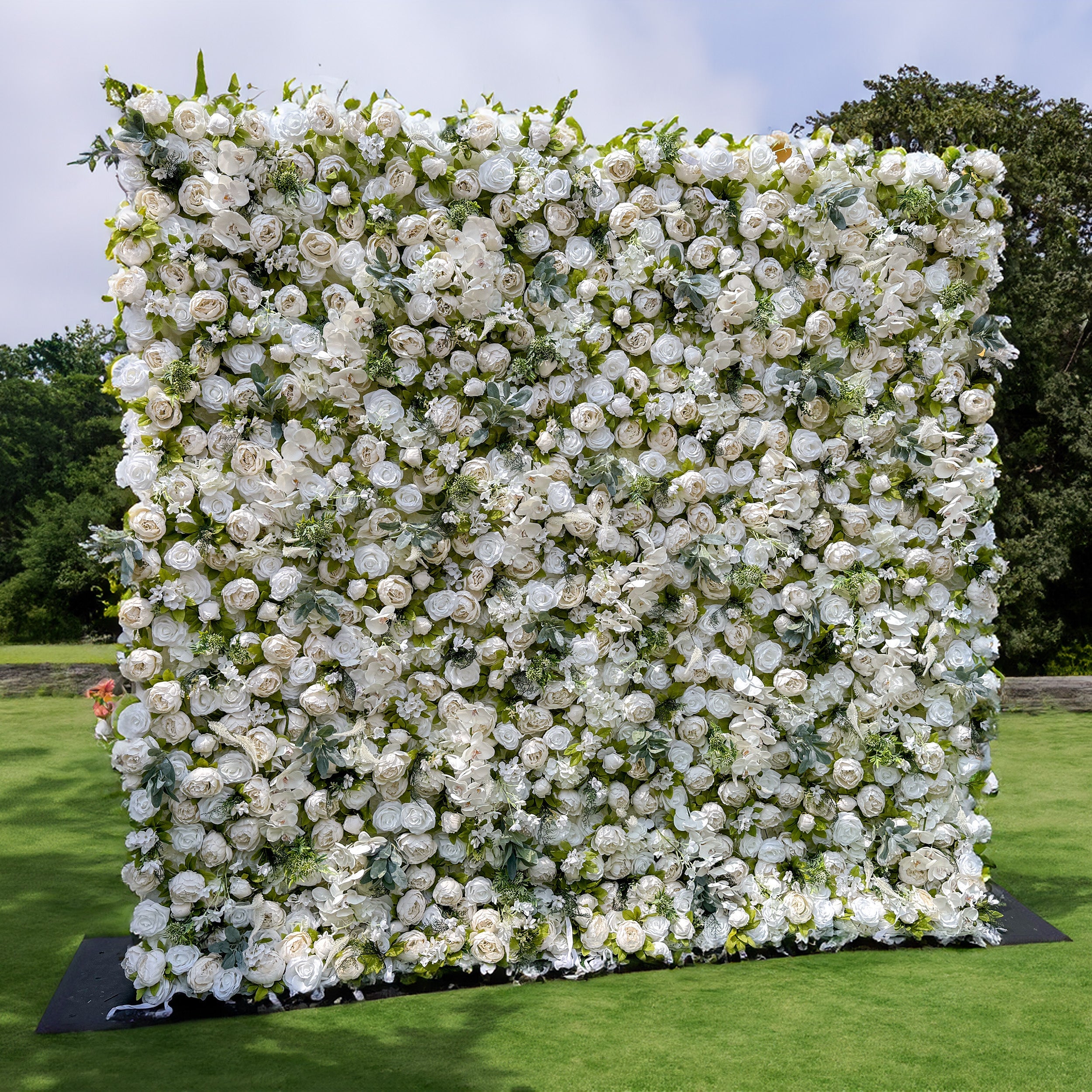 The image depicts a large, square - shaped flower wall. It is densely covered with white roses and green leaves, creating a lush and elegant appearance. The flower wall stands on a grassy area, with a backdrop of trees and greenery, enhancing its natural and serene setting. The arrangement of the flowers is uniform, giving the wall a full and luxurious look.