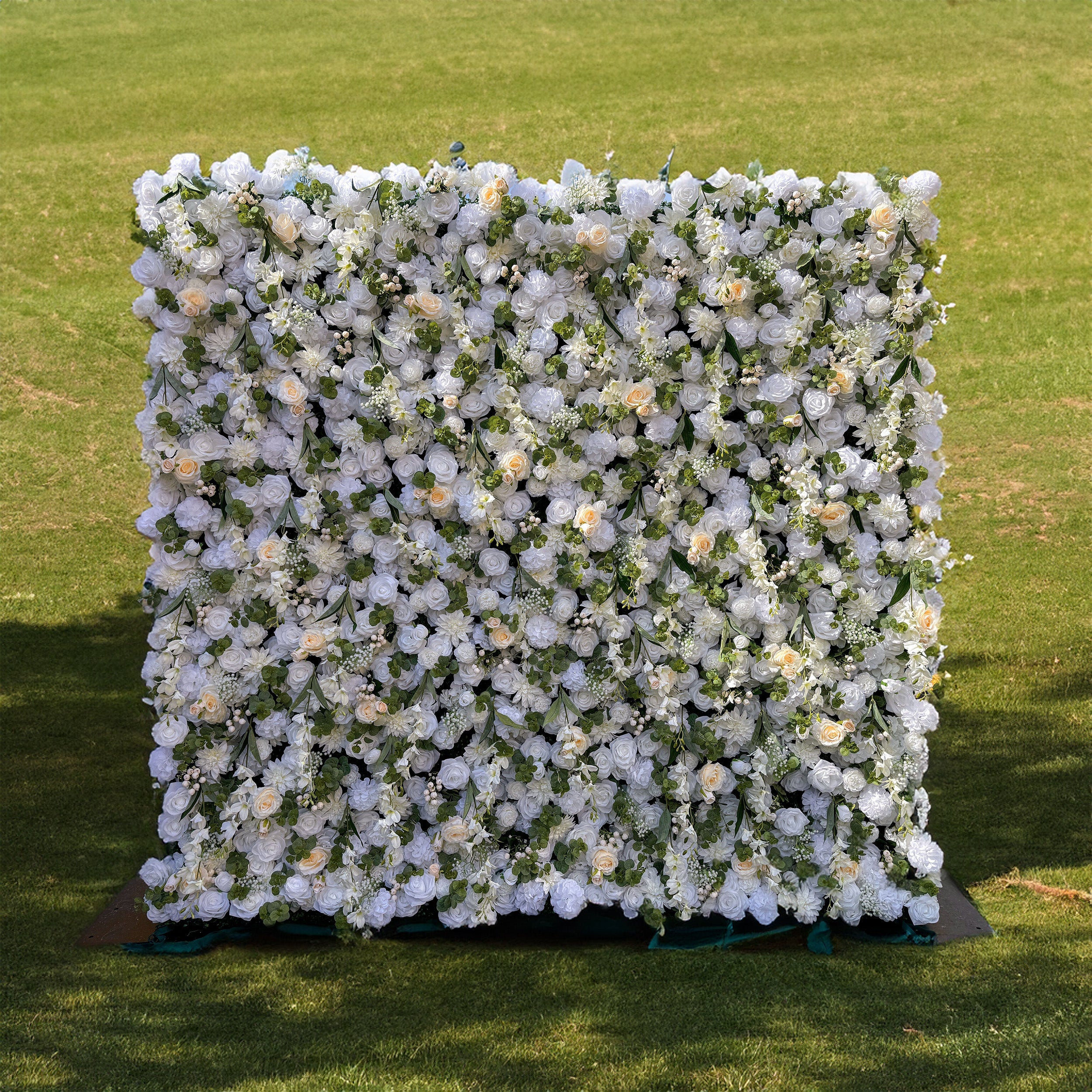 This is a beautiful artificial flower wall. It is densely covered with a variety of white flowers, including large, elegant roses and smaller blooms. Interspersed among the flowers are lush green leaves, creating a rich and textured appearance. The overall design is dense and full, giving a luxurious and abundant floral display, perfect for wedding or event backdrops.