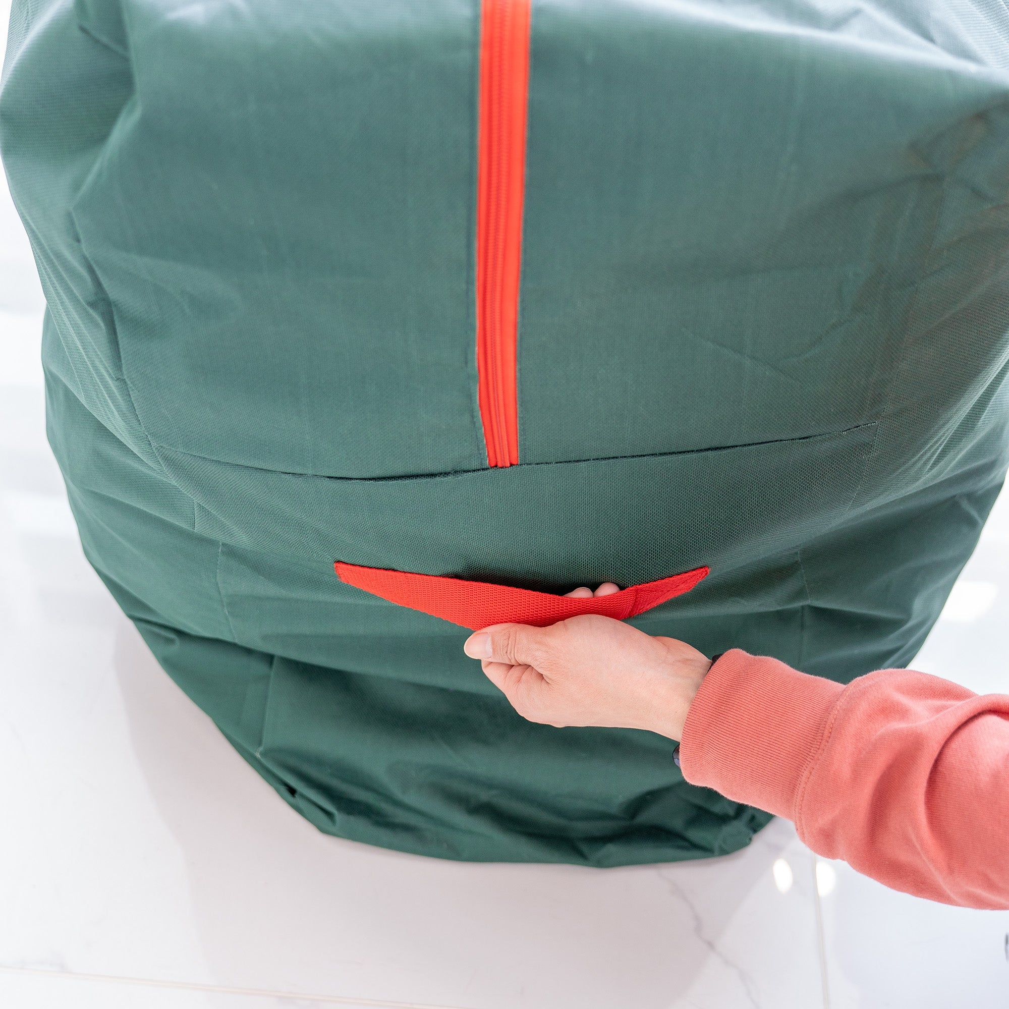 This image depicts the side of a green storage bag for flower walls. A vibrant red zipper runs vertically along the fabric, adding a striking contrast. A hand is seen gripping a red handle attached to the side of the bag, highlighting its functionality for carrying. The bag appears spacious and well - made, suitable for storing and transporting flower wall decorations. 