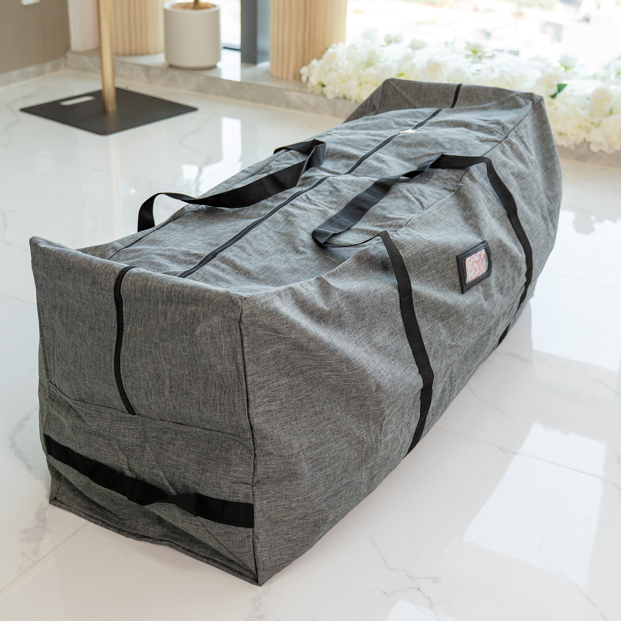 This image features a large, rectangular storage bag in a heathered gray color. The bag is equipped with sturdy black carrying straps and has a zipper running along its length, suggesting it can be opened wide for easy access. There is a small, rectangular label patch on the side of the bag. The bag is placed on a sleek, white marble - tiled floor, with a background that includes some white floral arrangements and modern, light - colored furniture elements, giving it a clean and contemporary setting. 