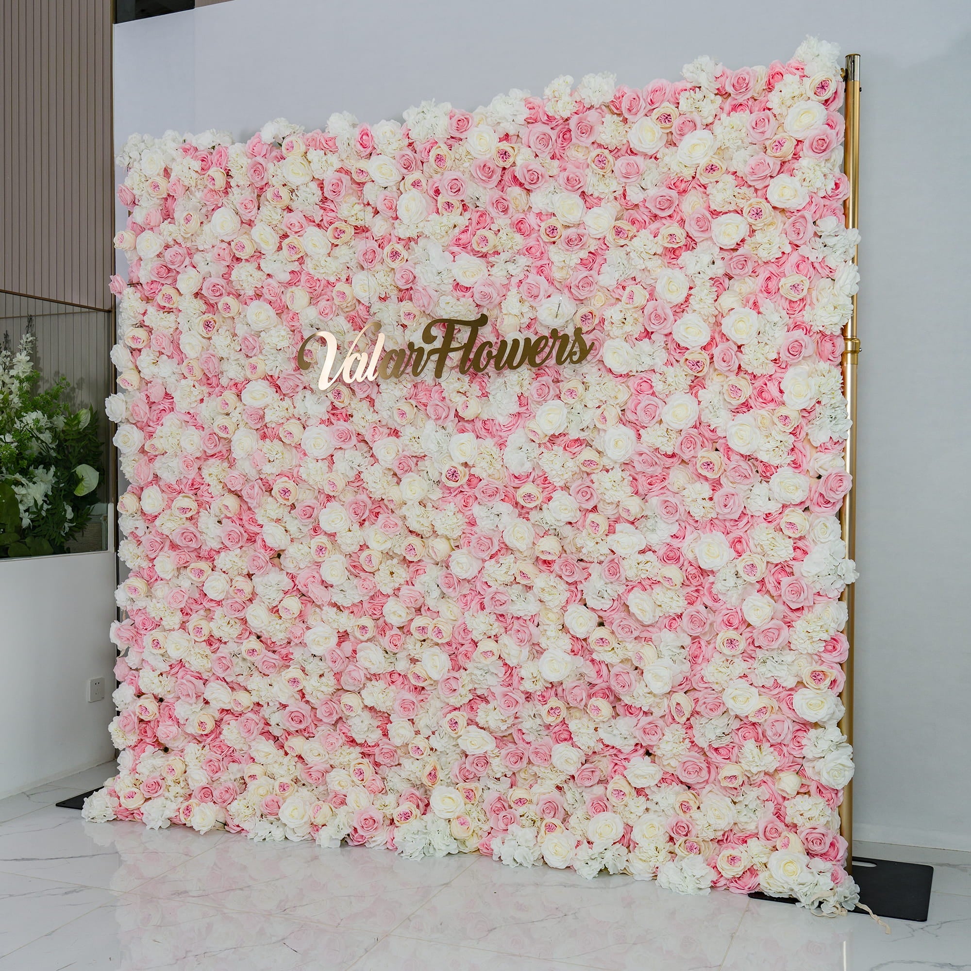 This photo showcases a lush floral backdrop densely adorned with pink and white roses. The soft pastel color palette creates a romantic and elegant atmosphere. The words "Valar Flowers" in elegant gold lettering stand out prominently. Ideal for weddings, bridal showers, or other romantic events, it exudes a dreamy and sophisticated style. 