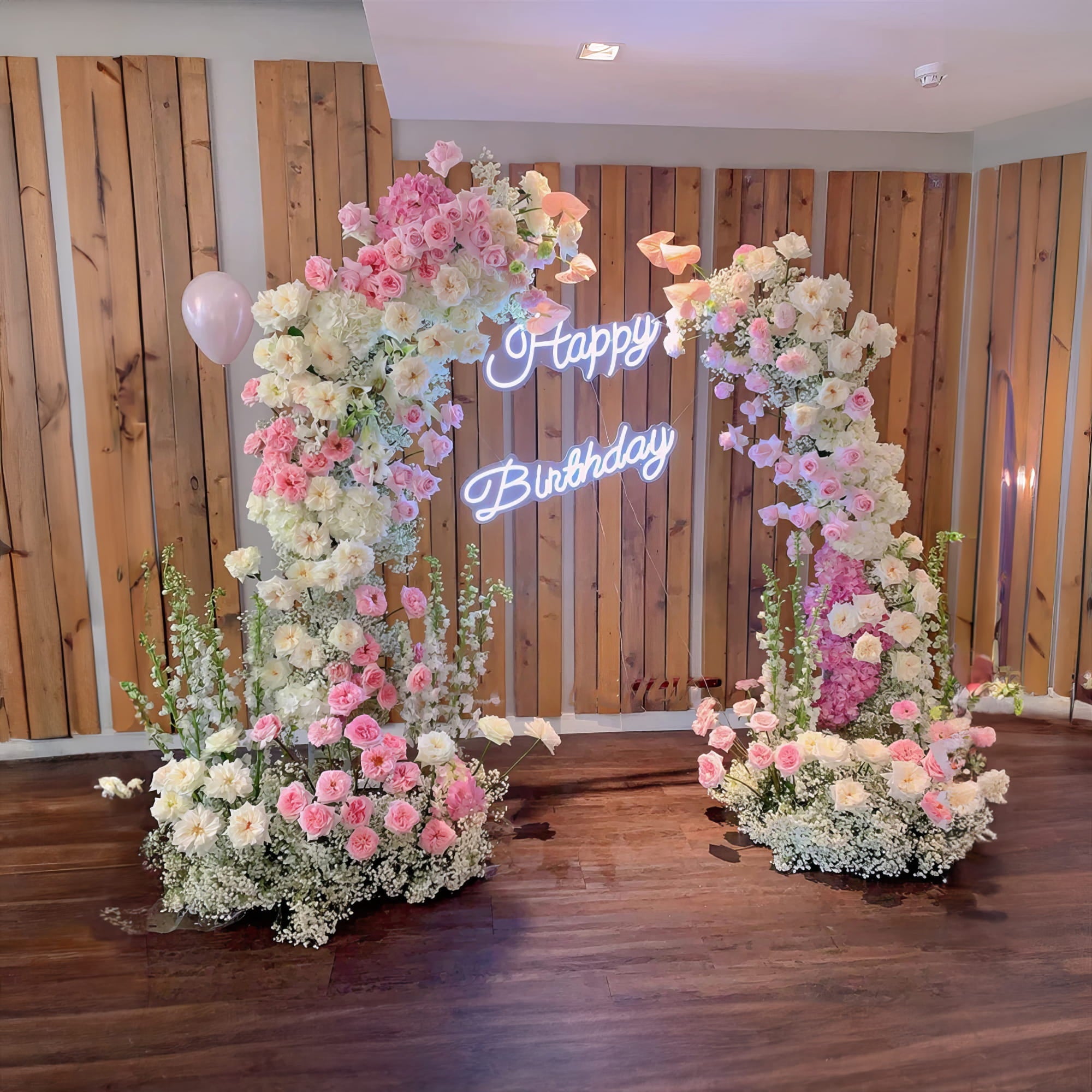 A charming floral arch with pink and white roses frames a neon "Happy Birthday" sign. Set against a wooden backdrop, it creates a festive and elegant ambiance, perfect for a birthday celebration.