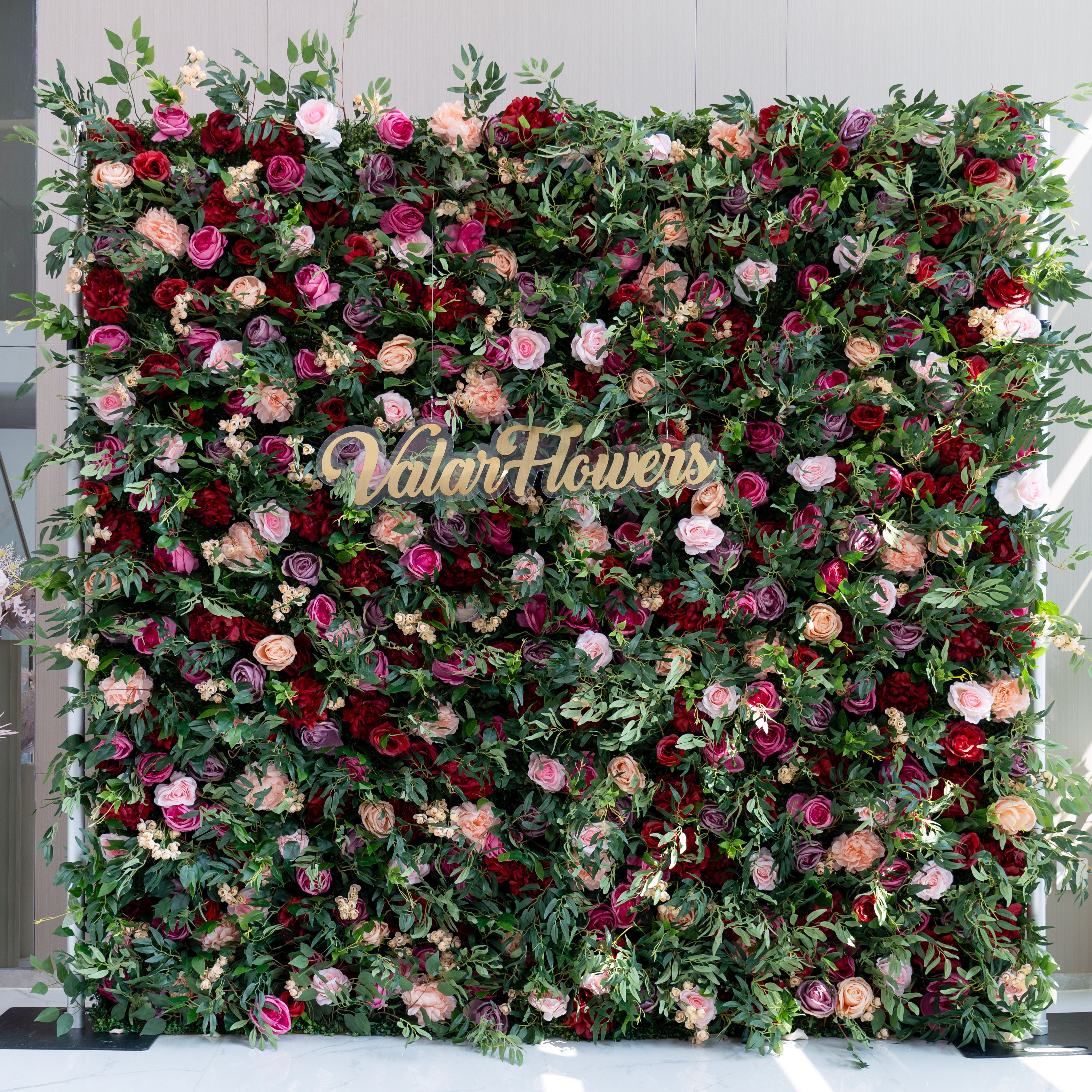 Vibrant artificial flower wall featuring a mix of red, pink, and blush roses interwoven with lush greenery, with the Valar Flowers logo displayed in the center.