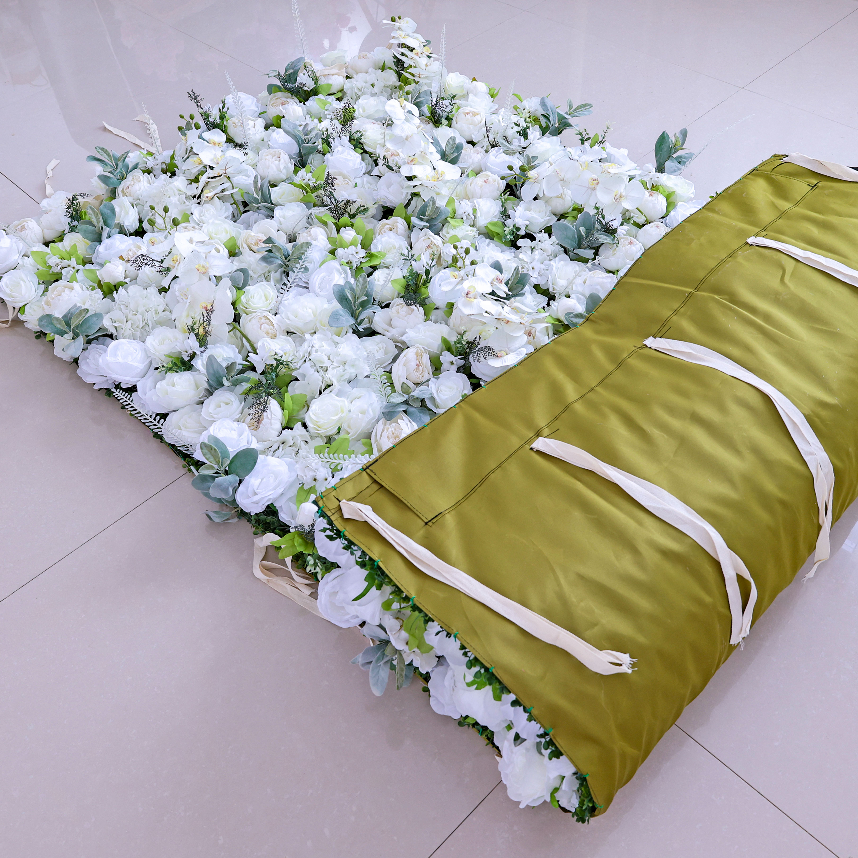 The image shows a ValarFlowers product: a rolled - up artificial flower wall with a green cover and white ties. It features a dense arrangement of white flowers, ideal for event decorations.