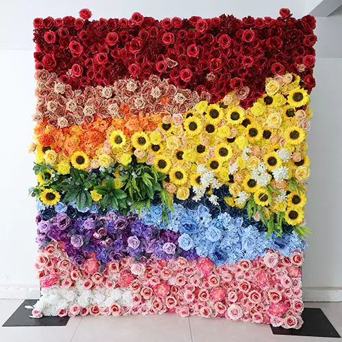 A vibrant flower wall, showcasing a medley of colors. It features sections of red roses, pastel flowers, bright sunflowers, lush green leaves, and a mix of blue and purple blossoms. Each segment of color blends seamlessly into the next, creating a captivating, multi-layered visual display. Set against a neutral wall, its brilliance stands out prominently.